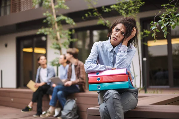 Young upset lady sitting on bench with colorful folders and thoughtfully looking aside while spending time in courtyard of university with students on background