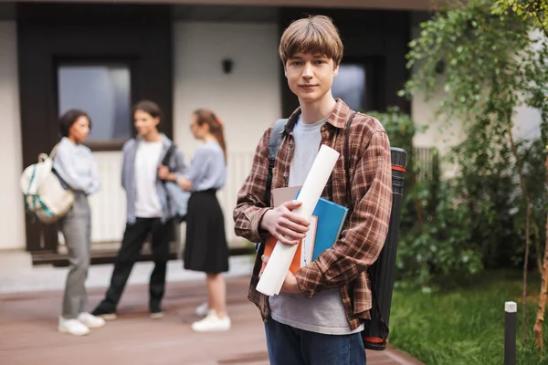 Portrait of young man standing with books in hands and dreamily looking in camera in courtyard of university with students on background