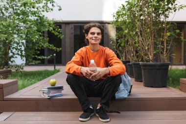 Şişe suyu el ve Kitaplar yakınındaki ve sersem bir kenara Üniversitesi avluda seyir ile oturan genç gülümseyen adam 