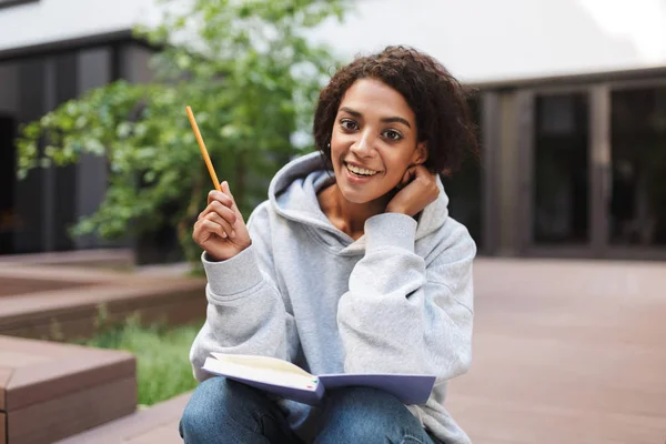 Pretty girl with dark curly hair sitting with open book on knees and happily looking in camera while studying in courtyard of university