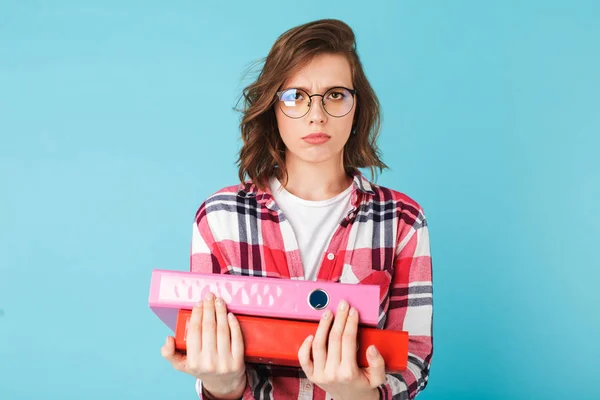 Young lady in eyeglasses and shirt standing with folders in hands and sadly looking in camera on over pink background