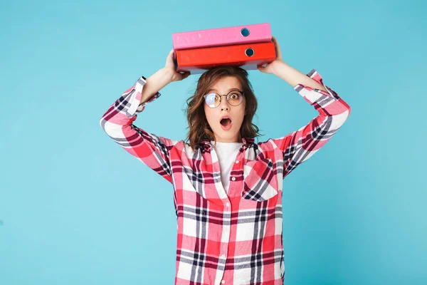 Young lady in eyeglasses and hipster shirt holding folders on head while surprised and amazedly looking in camera on over blue background