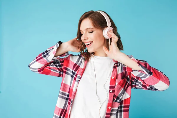 Portrait of young pretty lady in shirt dancing while listening music in headphones on over pink background