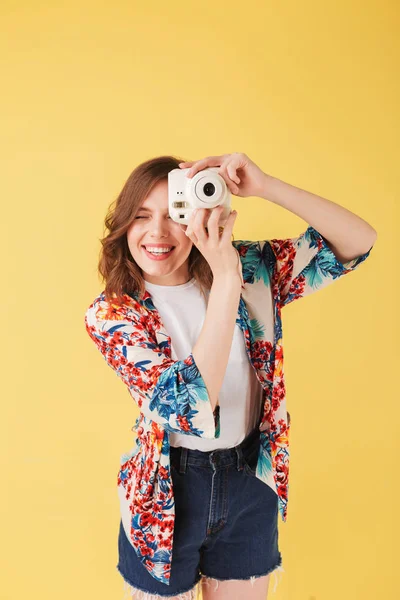 Portrait of pretty smiling lady in colorful shirt standing with little white camera on over pink background