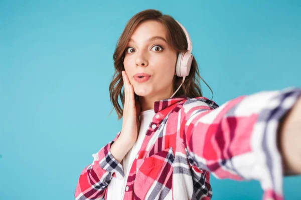 Portrait of young pretty lady in shirt listening music in headphones and surprisedly looking in camera on over pink background