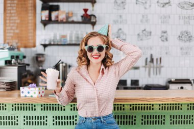 Gülümseyen güzel Bayan güneş gözlüğü ve barda oturan Doğum günü şapkası kamera seyir kafede elinde ve mutlu bir şekilde milkshake ile karşı
