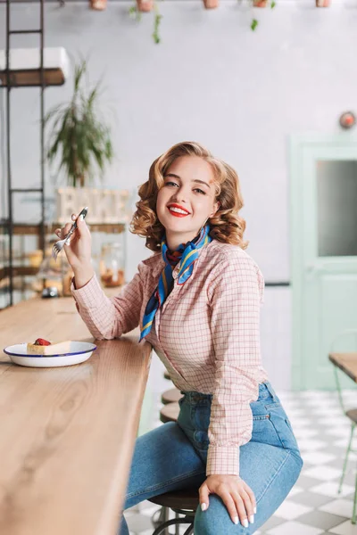 Smiling lady in shirt and jeans sitting at the bar counter in cafe with fork in hand and cake near and joyfully looking in camera