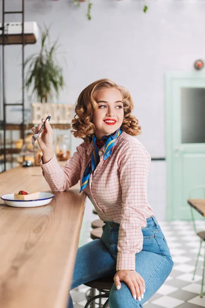 Pretty smiling lady in shirt and jeans sitting at the bar counter in cafe with fork in hand and cake near and dreamily looking aside