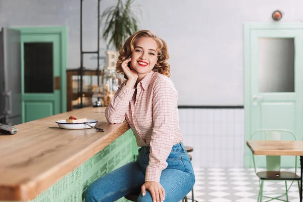 Beautiful lady in shirt and jeans sitting at the bar counter with piece of cake near and joyfully looking in camera while spending time in cafe