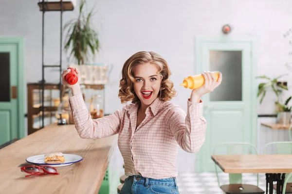 Pretty lady in shirt sitting with ketchup and mustard bottles in hands and joyfully looking in camera while spending time in cafe