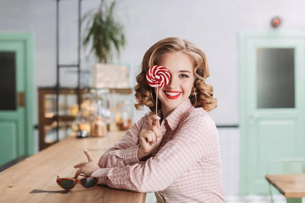 Young beautiful lady sitting at the bar counter and covering her eye with lollipop candy while happily looking in camera in cafe