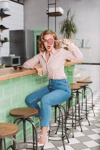 Young lady in shirt and jeans sitting at the bar counter and covering her eye with lollipop candy while amazedly looking in camera in cafe