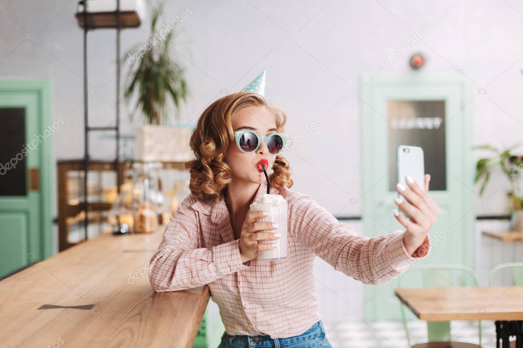 Beautiful girl in sunglasses and birthday cap sitting at the bar counter and drinking milkshake while taking cute photos on her cellphone in cafe