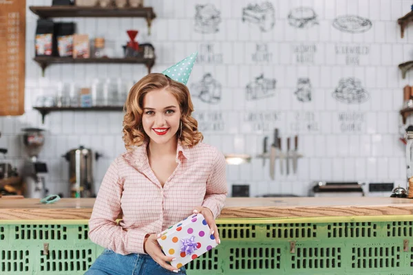 Young smiling lady in shirt and birthday cap sitting at the bar counter with present box in hands and joyfully looking in camera in cafe