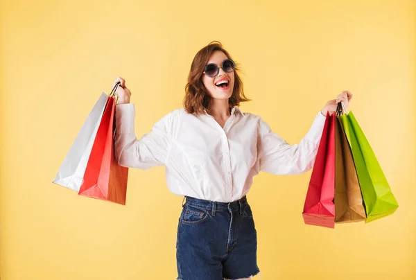 Portrait of young joyful lady in sunglasses standing with colorful shopping bags in hands on over pink background. Happy woman standing in white shirt and denim shorts