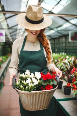 Önlük ve ayakta ve serada çalışırken metal sepet çiçek holding şapkasını gülümseyen çiçekçi