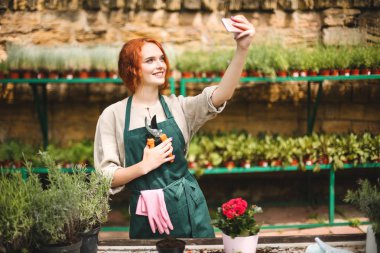 Bahçe makası ve sera ön cep telefonu kamera çekimi fotoğrafları ile önlük ayakta genç gülümseyen çiçekçi