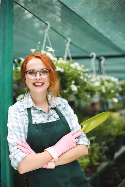 Küçük bahçe kürekle el ayakta önlük ve pembe eldiven güzel çiçekçi. Gözlük sevinçle serada çalışan genç gülümseyen kadın