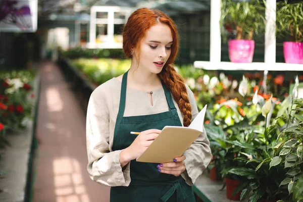 Serada çalışırken Not Defteri'nde yazma el ve düşünceli kalem ile önlük ayakta genç çiçekçi
