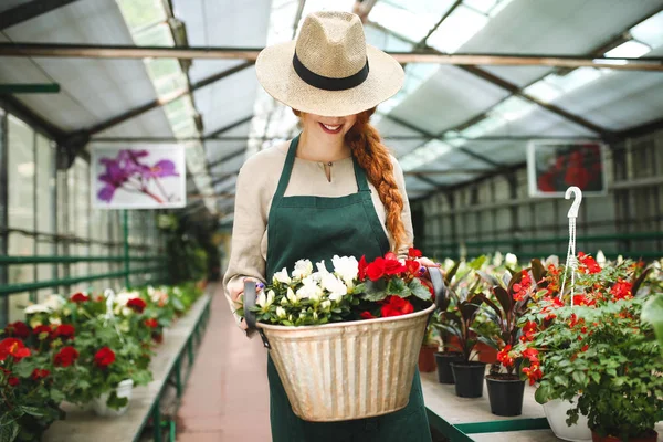 Smiling florist in apron standing and holding flowers in metal basket while covering her face with hat in greenhouse