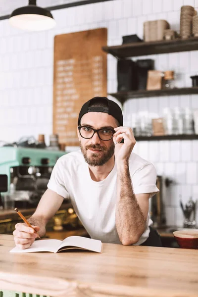 Smiling Man Eyeglasses Cap Standing Bar Counter Pencil Notepad Happily — Stock Photo, Image