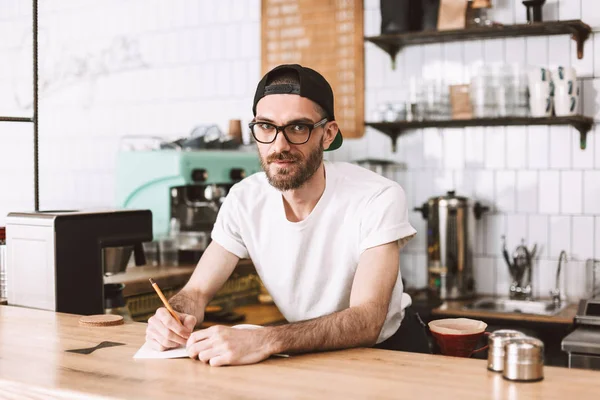 Smiling man in eyeglasses and cap standing behind bar counter with pencil and notepad and dreamily looking in camera in cafe