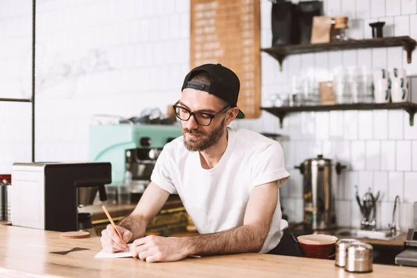 Thoughtful Man Eyeglasses Cap Standing Bar Counter Writing Notepad While — Stock Photo, Image