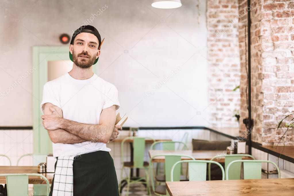 Young man in black cap,white t-shirt and apron standing and dreamily looking in camera while working in cafe