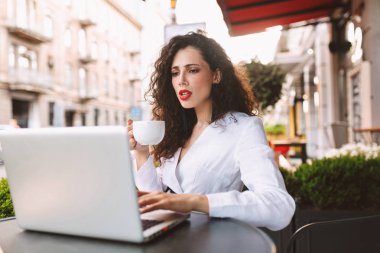 Beyaz kostüm ile el ve düşünceli sokakta kafede dizüstü kullanarak kahve masada oturan siyah kıvırcık saçlı Genç Bayan