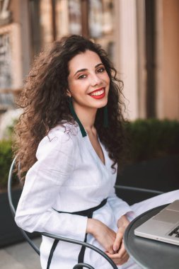 Masada oturan ve sevinçle kamera sokakta vakit kafede süre seyir beyaz kostüm siyah kıvırcık saçlı genç gülümseyen kadın 