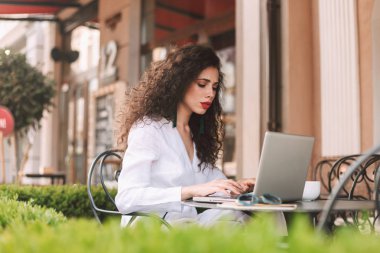 Masada oturan ve düşünceli sokakta kafede dizüstü bilgisayar üzerinde çalışan beyaz kostüm siyah kıvırcık saçlı Genç Bayan 