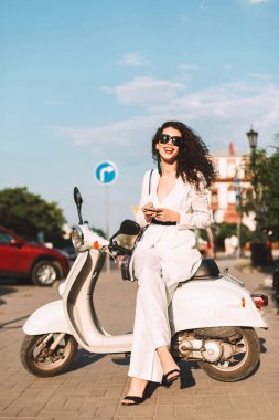 Siyah kıvırcık saçlı beyaz kostüm ve beyaz motosiklet üzerinde oturan ve mutlu bir şekilde fotoğraf makinesindeki Caddesi üzerinde arka plan üzerinde şehir manzaralı seyir güneş gözlüğü ile gülümseyen güzel Bayan