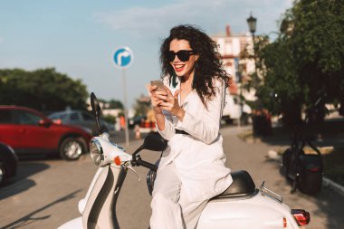 Siyah kıvırcık saçlı beyaz kostüm ve beyaz motosiklet üzerinde oturan ve mutlu bir şekilde arka planda şehir manzaralı sokakta cep telefonunu kullanarak güneş gözlüğü ile neşeli bayan