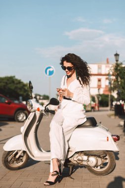 Beyaz kostüm ve beyaz motosiklet üzerinde oturan ve arka plan üzerinde şehir manzaralı Caddesi üzerinde cep telefonu kullanarak saunglasses siyah kıvırcık saçlı hoş gülümseyen kadın