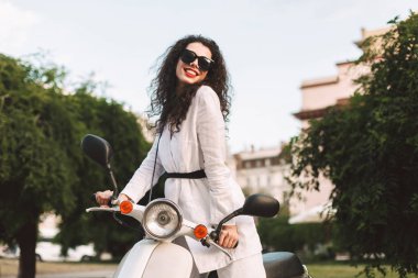 Beyaz kostüm ve beyaz motosiklet üzerinde oturan ve mutlu bir şekilde kamera seyir sokağa güneş gözlüğü siyah kıvırcık saçlı genç gülümseyen kadın