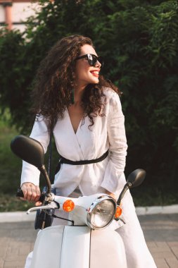 Siyah kıvırcık saçlı beyaz kostüm ve beyaz motosiklet üzerinde oturan ve mutlu bir kenara Caddesi üzerinde seyir güneş gözlüğü ile gülümseyen güzel Bayan