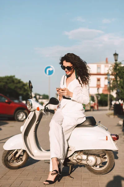 Beyaz kostüm ve beyaz motosiklet üzerinde oturan ve arka plan üzerinde şehir manzaralı Caddesi üzerinde cep telefonu kullanarak saunglasses siyah kıvırcık saçlı hoş gülümseyen kadın