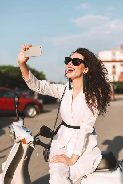 年轻开朗的妇女与深色卷发的白色服装和太阳镜坐在白色的脚踏车 并愉快地拍摄手机正面相机的照片 而在城市街道上花时间 — 图库照片