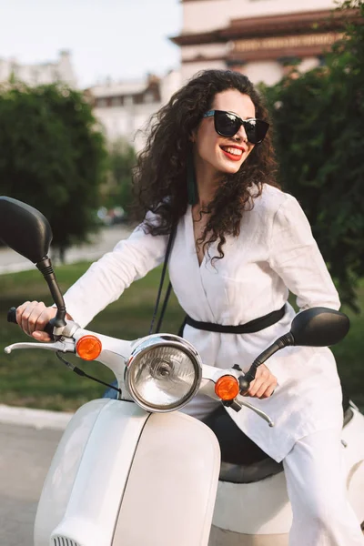 Siyah kıvırcık saçlı beyaz kostüm ve beyaz motosiklet üzerinde oturan ve sevinçle bir kenara Caddesi üzerinde seyir güneş gözlüğü ile oldukça gülümseyen kadın