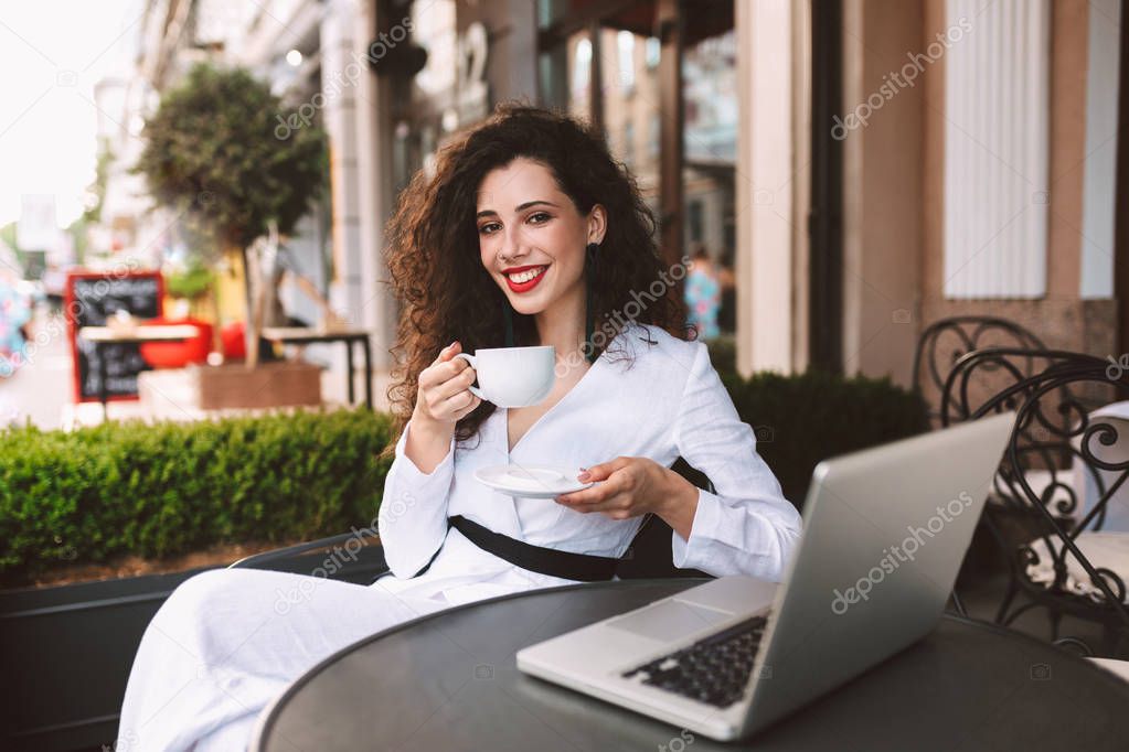 Beautiful smiling lady with dark curly hair in white costume sitting at the table with laptop and cup of coffee in hands while happily looking in camera in cafe on street