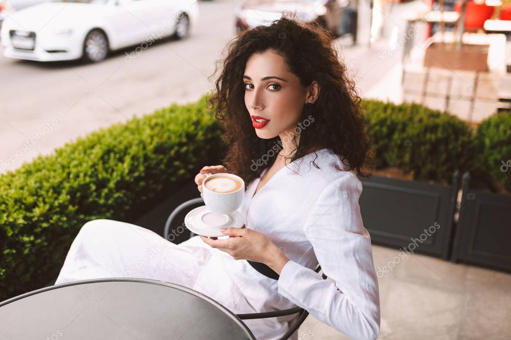 Beautiful lady with dark curly hair in white costume sitting at the table with cup of coffee in hands and thoughtfully looking in camera in cafe on street