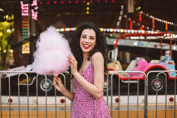 Joyful lady with dark curly hair in dress standing with pink cotton candy in hand and happily looking in camera while spending time in amusement park