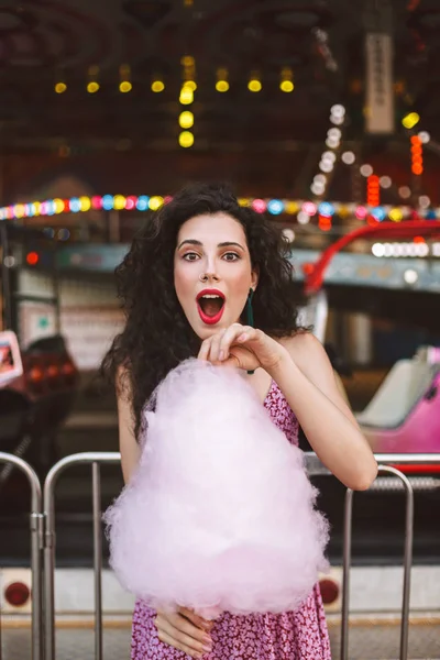 Elbise ayakta pembe pamuk şeker el ve amazedly eğlence parkı olan kamera seyir ile siyah kıvırcık saçlı Genç Bayan