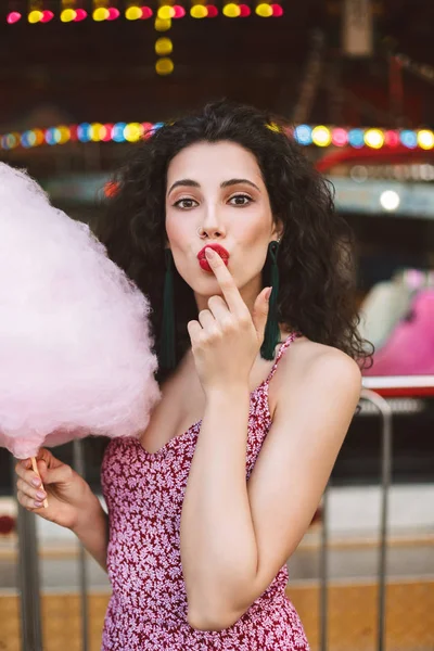 Genç güzel bayan elbise ayakta pembe pamuk şeker kamera seyir eğlence parkında el ve sersem ile siyah kıvırcık saçlı