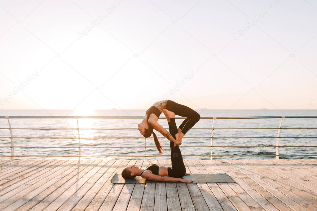 Photo of two beautiful ladies in black sporty tops and leggings training yoga poses by the sea. Young women practicing yoga together with beautiful sea view on background