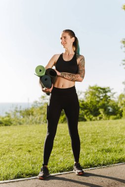 Genç gülümseyen Bayan siyah spor top ve yoga paspaslar elinde ile ayakta ve sersem bir kenara park seyir tozluk