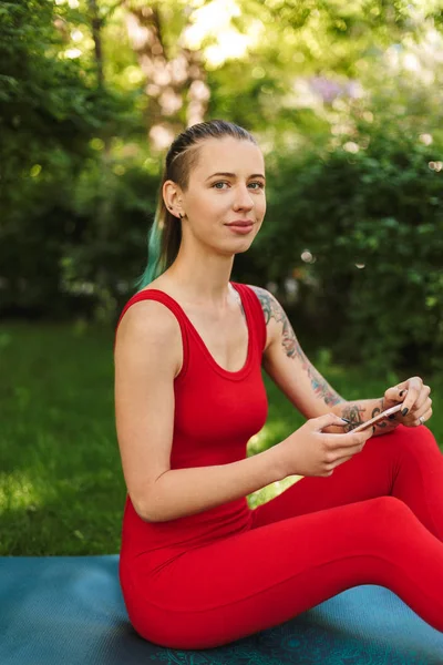 美丽的女人在红色的连身裤坐在瑜伽垫上的手机和朦胧地看着相机 而在公园练习瑜伽 — 图库照片