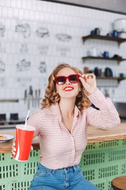 Güneş gözlüğü ve barda oturan gömlek gülümseyen güzel Bayan soda kamera seyir kafede elinde ve mutlu bir şekilde su ile karşı