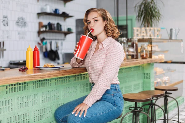 Young beautiful lady in shirt and jeans sitting at the bar counter and drinking soda water while dremily looking in camera in cafe