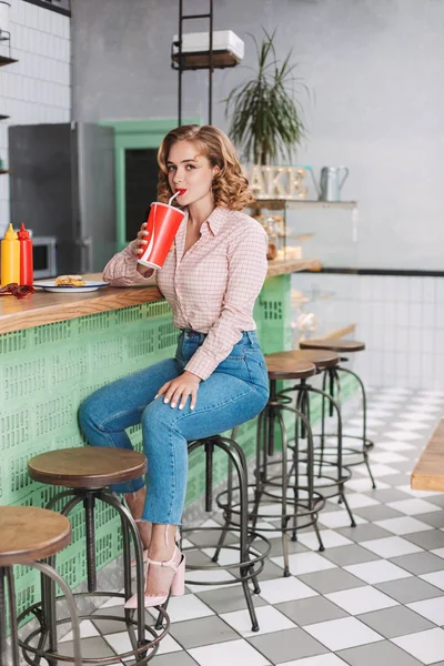 Pretty lady in shirt and jeans sitting at the bar counter and drinking soda water while happily looking in camera in cafe